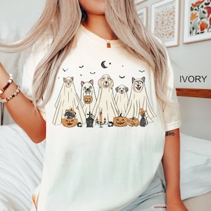 Peut inclure: Un t-shirt blanc avec une illustration vintage de cinq chiens habill&eacute;s en fant&ocirc;mes pour Halloween. Les chiens tiennent des citrouilles et il y a des chauves-souris et une lune croissante en arri&egrave;re-plan. Le texte "IVORY" est dans le coin sup&eacute;rieur droit de l'image.
