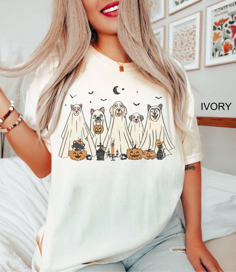 Peut inclure: Un t-shirt blanc avec une illustration vintage de cinq chiens habill&eacute;s en fant&ocirc;mes pour Halloween. Les chiens tiennent des citrouilles et il y a des chauves-souris et une lune croissante en arri&egrave;re-plan. Le texte "IVORY" est dans le coin sup&eacute;rieur droit de l'image.
