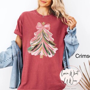 Op de afbeelding: Karmizijnrood T-shirt met een kerstboomontwerp. De boom is versierd met roze, groene en gouden penseelstreken, een ster en een strik. Het shirt heeft opgerolde mouwen en komt uit de Etsy shop Lovin What U Wear.