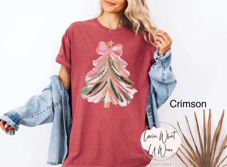 Op de afbeelding: Karmizijnrood T-shirt met een kerstboomontwerp. De boom is versierd met roze, groene en gouden penseelstreken, een ster en een strik. Het shirt heeft opgerolde mouwen en komt uit de Etsy shop Lovin What U Wear.