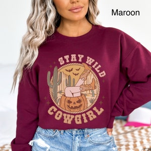 May include: A maroon sweatshirt featuring a graphic print of a woman in cowboy boots sitting in a jack-o-lantern. The design also includes cacti, bats, and the text "Stay Wild Cowgirl".
