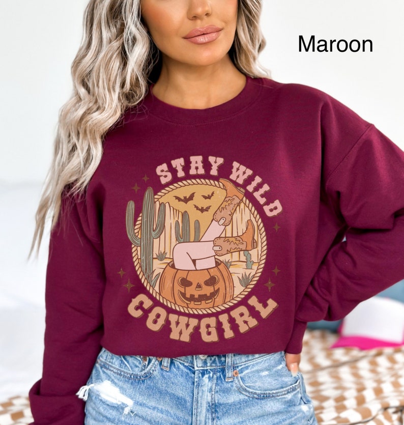 May include: A maroon sweatshirt featuring a graphic print of a woman in cowboy boots sitting in a jack-o-lantern. The design also includes cacti, bats, and the text "Stay Wild Cowgirl".