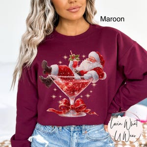 May include: Maroon sweatshirt featuring a festive graphic of Santa Claus relaxing in a martini glass filled with red glitter. The design includes a red bow and the text "Lovin What U Wear".