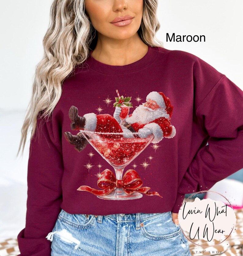 May include: Maroon sweatshirt featuring a festive graphic of Santa Claus relaxing in a martini glass filled with red glitter. The design includes a red bow and the text "Lovin What U Wear".