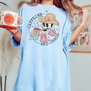 Pu&ograve; includere: Una maglietta blu chiaro con una grafica di uno scheletro che indossa un cappello da cowboy e tiene una tazza di caff&egrave;. Il testo "Caffeine and Chaos" &egrave; scritto intorno allo scheletro.