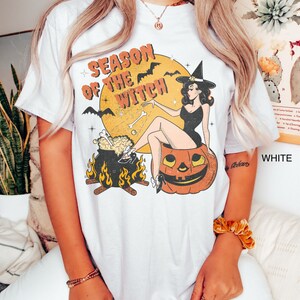 Puede incluir: Camiseta blanca con una ilustraci&oacute;n vintage de una bruja sentada en una calabaza. La bruja sostiene un hueso y un caldero est&aacute; frente a ella. El texto "Season of the Witch" est&aacute; impreso en la camiseta.