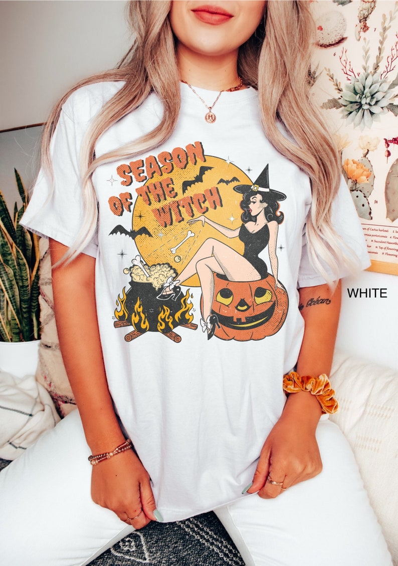 Puede incluir: Camiseta blanca con una ilustraci&oacute;n vintage de una bruja sentada en una calabaza. La bruja sostiene un hueso y un caldero est&aacute; frente a ella. El texto "Season of the Witch" est&aacute; impreso en la camiseta.