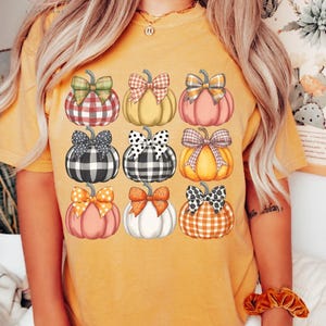Peut inclure: T-shirt moutarde avec une grille de neuf citrouilles avec des n&oelig;uds &agrave; motifs vari&eacute;s. Les citrouilles sont dans les tons de rose, orange, jaune et blanc. Le texte "Lovin What U Wear" est en haut &agrave; gauche.
