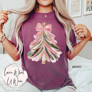 Op de afbeelding: Bessenkleurig T-shirt met een kerstboomontwerp. De boom is versierd met roze, groene en gouden penseelstreken, bekroond met een roze strik en een gouden ster. De tekst "Lovin What U Wear" staat linksonder.
