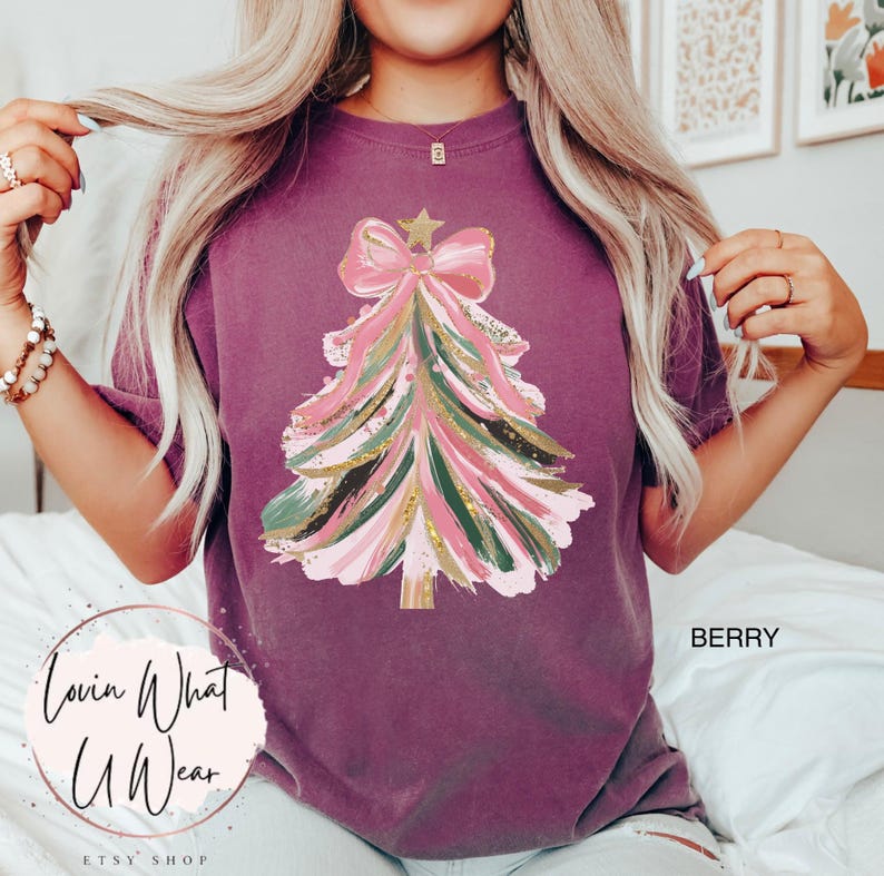 Op de afbeelding: Bessenkleurig T-shirt met een kerstboomontwerp. De boom is versierd met roze, groene en gouden penseelstreken, bekroond met een roze strik en een gouden ster. De tekst "Lovin What U Wear" staat linksonder.