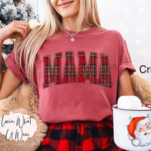 May include: Crimson-colored t-shirt with the word "MAMA" in a plaid design. The person is holding a mug with a Santa Claus graphic. The shirt is paired with red and black plaid pajama pants.