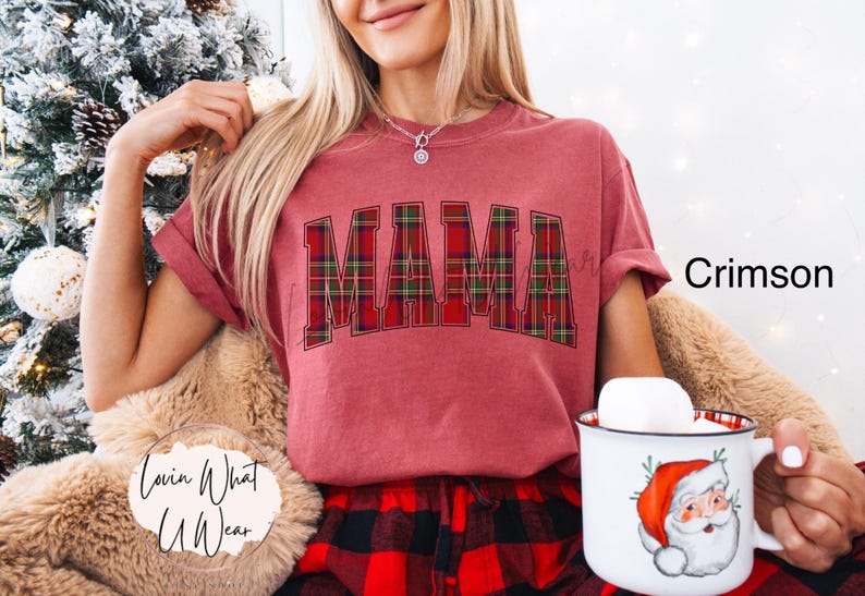 May include: Crimson-colored t-shirt with the word "MAMA" in a plaid design. The person is holding a mug with a Santa Claus graphic. The shirt is paired with red and black plaid pajama pants.