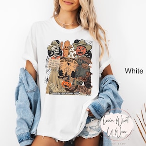 May include: White t-shirt with a Halloween-themed graphic. The design features a skeleton, cactus, and pumpkin wearing cowboy hats, along with the text "October 31" and "Halloween Never Die". The shirt also has a spider web and a ghost.