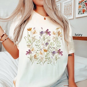 May include: A white t-shirt with a floral print featuring a variety of flowers in shades of purple, yellow, and green. The text "Ivory" is visible in the bottom right corner of the image.