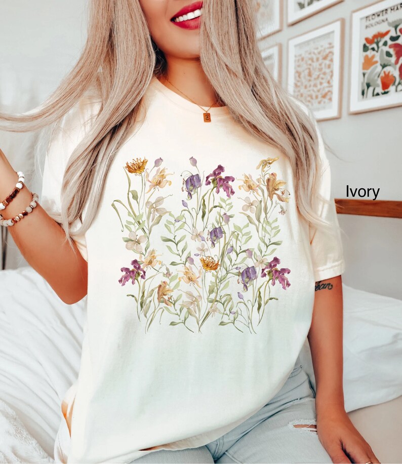 May include: A white t-shirt with a floral print featuring a variety of flowers in shades of purple, yellow, and green. The text "Ivory" is visible in the bottom right corner of the image.