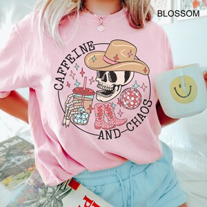 Pu&ograve; includere: Maglietta rosa con una grafica di uno scheletro che indossa un cappello da cowboy, tiene una tazza di caff&egrave; e una palla da discoteca. Il testo "Caffeine and Chaos" &egrave; scritto intorno alla grafica.