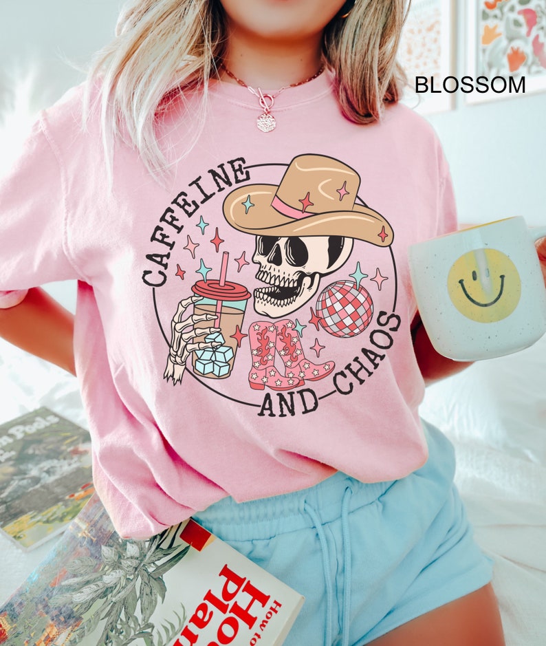 Pu&ograve; includere: Maglietta rosa con una grafica di uno scheletro che indossa un cappello da cowboy, tiene una tazza di caff&egrave; e una palla da discoteca. Il testo "Caffeine and Chaos" &egrave; scritto intorno alla grafica.