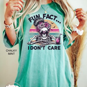 May include: A turquoise blue t-shirt with a graphic of a skeleton wearing sunglasses and a flower crown. The graphic is set against a sunset background with the text "Fun Fact... I Don't Care".