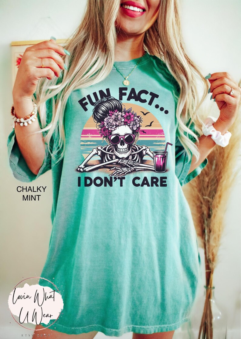 May include: A turquoise blue t-shirt with a graphic of a skeleton wearing sunglasses and a flower crown. The graphic is set against a sunset background with the text "Fun Fact... I Don't Care".