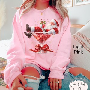 May include: Light pink sweatshirt featuring a graphic of Santa Claus relaxing in a martini glass filled with red glitter. The martini glass has a red bow at the base. The text "Lovin What U Wear" is on the lower right.