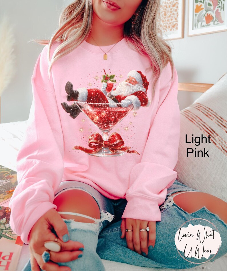 May include: Light pink sweatshirt featuring a graphic of Santa Claus relaxing in a martini glass filled with red glitter. The martini glass has a red bow at the base. The text "Lovin What U Wear" is on the lower right.