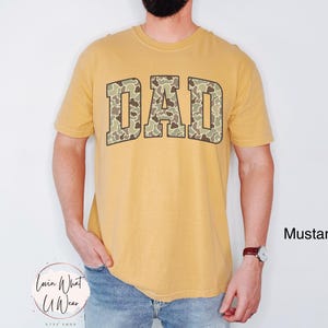 May include: Mustard-colored t-shirt with the word "DAD" in a camouflage pattern across the chest. The shirt is short-sleeved and has a crew neck. The text "Mustard" is in the lower right corner.