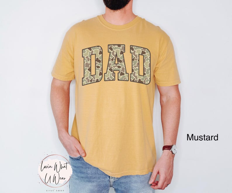 May include: Mustard-colored t-shirt with the word "DAD" in a camouflage pattern across the chest. The shirt is short-sleeved and has a crew neck. The text "Mustard" is in the lower right corner.
