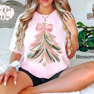 Op de afbeelding: Lichtroze T-shirt met een kerstboomontwerp met roze, groene en gouden accenten. De boom is versierd met een grote roze strik en een ster. Het woord "Blossom" staat rechtsonder.