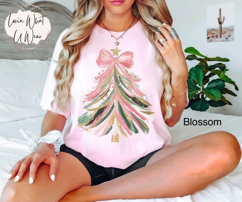 Op de afbeelding: Lichtroze T-shirt met een kerstboomontwerp met roze, groene en gouden accenten. De boom is versierd met een grote roze strik en een ster. Het woord "Blossom" staat rechtsonder.