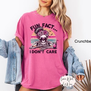 May include: Pink t-shirt with a graphic of a skeleton wearing a flower crown and sunglasses, sitting with a drink. The text "Fun Fact... I Don't Care" is printed on the shirt.