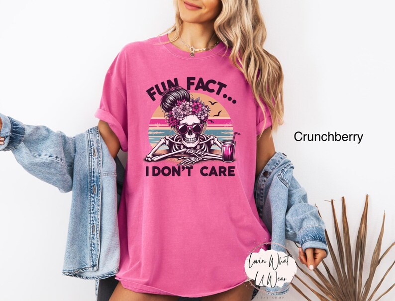 May include: Pink t-shirt with a graphic of a skeleton wearing a flower crown and sunglasses, sitting with a drink. The text "Fun Fact... I Don't Care" is printed on the shirt.