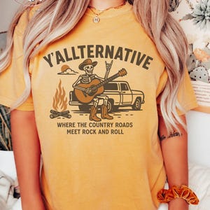 May include: Mustard-colored t-shirt with the text "Y'allternative" and a graphic of a skeleton playing guitar in front of a truck. The design includes a campfire and the phrase "Where the country roads meet rock and roll."