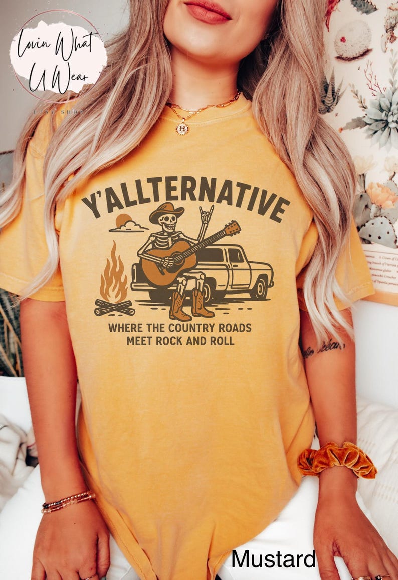 May include: Mustard-colored t-shirt with the text "Y'allternative" and a graphic of a skeleton playing guitar in front of a truck. The design includes a campfire and the phrase "Where the country roads meet rock and roll."