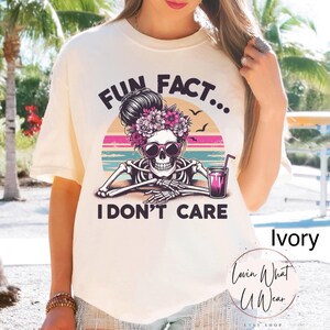 May include: Ivory t-shirt with a graphic of a skeleton with flowers in its hair, holding a glass of pink liquid. The text "Fun Fact... I Don't Care" is printed on the shirt.