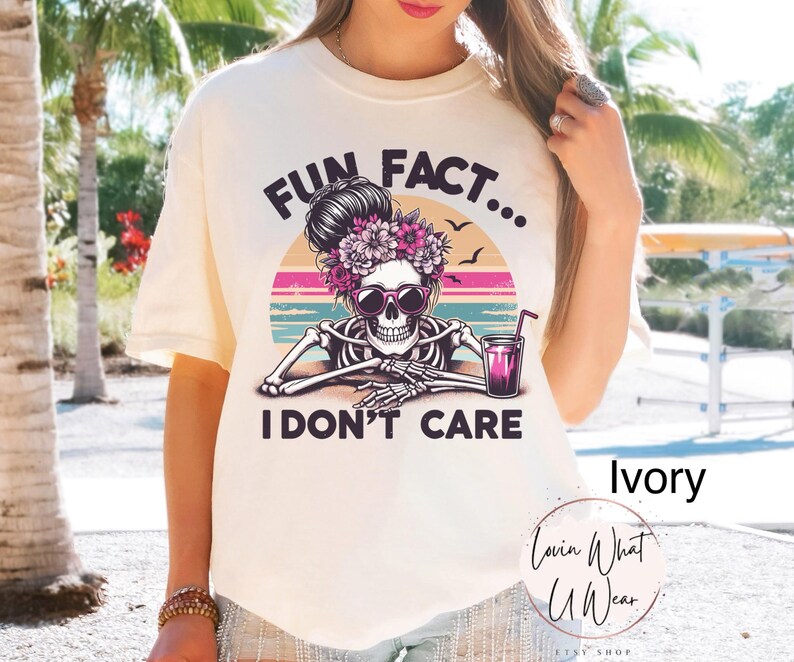 May include: Ivory t-shirt with a graphic of a skeleton with flowers in its hair, holding a glass of pink liquid. The text "Fun Fact... I Don't Care" is printed on the shirt.