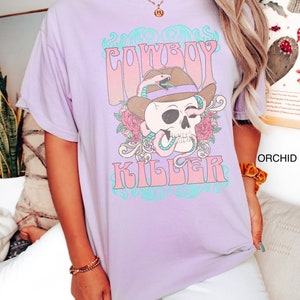 Op de afbeelding: Een lavendelkleurig T-shirt met een grafische print van een schedel die een cowboyhoed en een slang draagt. De tekst "COWBOY KILLER" is gedrukt in roze en blauwe letters.