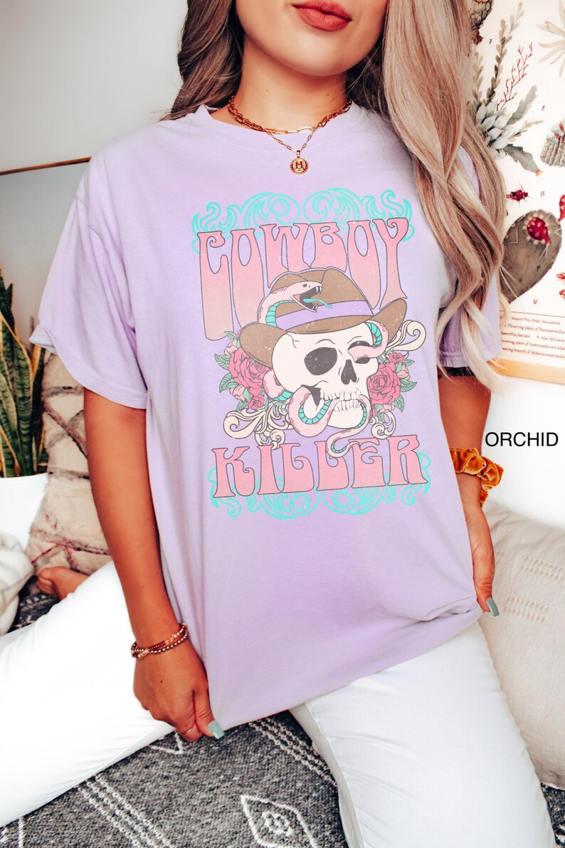 Op de afbeelding: Een lavendelkleurig T-shirt met een grafische print van een schedel die een cowboyhoed en een slang draagt. De tekst "COWBOY KILLER" is gedrukt in roze en blauwe letters.