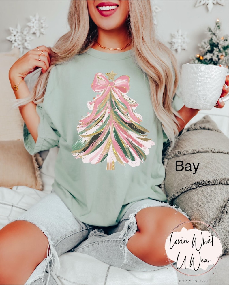 Op de afbeelding: Saliegroen T-shirt met een kerstboomontwerp met roze, gouden en groene penseelstreken. De boom is versierd met een roze strik en een gouden ster. Het shirt heeft een relaxte pasvorm en wordt gedragen met een spijkerbroekshort.