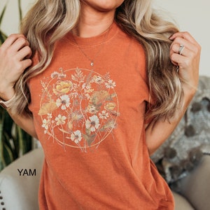 May include: A burnt orange t-shirt featuring a floral print in shades of yellow, white, and brown. The floral print is enclosed within a circle with a gold outline.