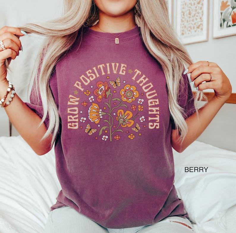 Op de afbeelding: Een paars T-shirt met een grafisch ontwerp van bloemen en vlinders. De tekst "Grow Positive Thoughts" staat in een cirkel rond de bloemen.