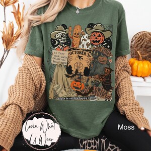 May include: Moss green t-shirt with a Halloween-themed graphic. The design includes a skeleton, jack-o'-lantern, and the text "October 31". Additional text reads "Is that your ghost or are you real here?" and "Halloween will never die".
