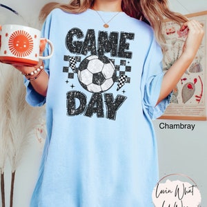 May include: Light blue t-shirt with the words "GAME DAY" in a distressed black font, a soccer ball, and checkered flags. The shirt is a relaxed fit and has short sleeves. The shirt is made of a soft material.
