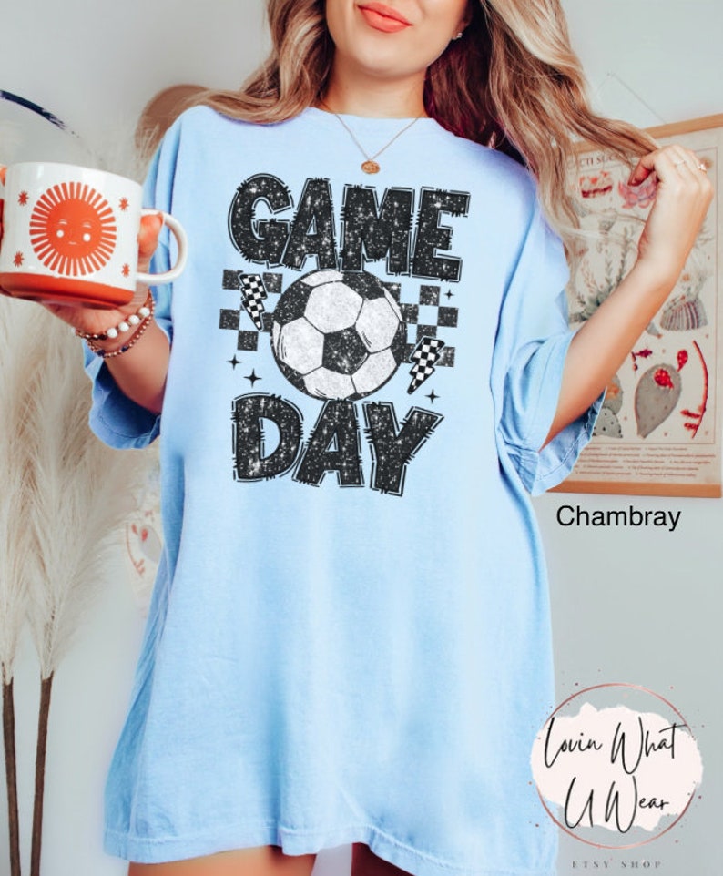 May include: Light blue t-shirt with the words "GAME DAY" in a distressed black font, a soccer ball, and checkered flags. The shirt is a relaxed fit and has short sleeves. The shirt is made of a soft material.