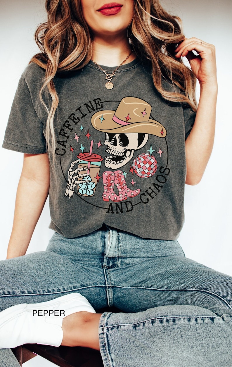 Pu&ograve; includere: Una maglietta grigia con una grafica di uno scheletro che indossa un cappello da cowboy e tiene una tazza di caff&egrave;. Il testo "CAFFEINE AND CHAOS" &egrave; scritto intorno allo scheletro.