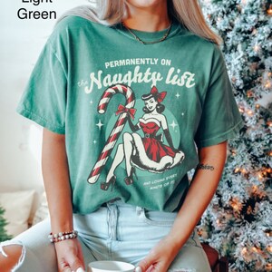 May include: A light green t-shirt with a vintage-style graphic of a woman in a red dress holding a candy cane. The text on the shirt reads "Permanently on the Naughty List and loving every minute of it!"