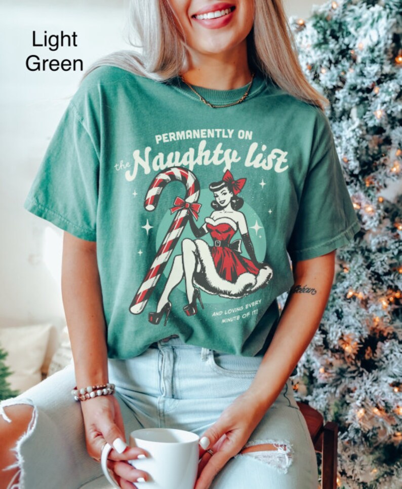 May include: A light green t-shirt with a vintage-style graphic of a woman in a red dress holding a candy cane. The text on the shirt reads "Permanently on the Naughty List and loving every minute of it!"