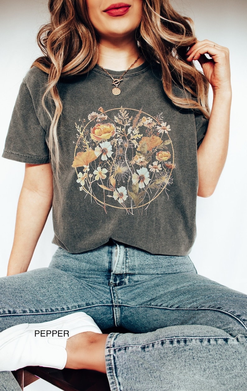 May include: A dark grey t-shirt featuring a floral print in shades of yellow, white, and brown. The flowers are arranged in a circle with a gold outline. The shirt is being worn by a person with long blonde hair and a gold necklace.