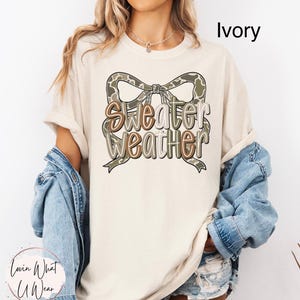 May include: Ivory-colored t-shirt with a bow-shaped graphic in shades of brown and green, featuring the words "Sweater Weather". The shirt is part of a casual wear collection.