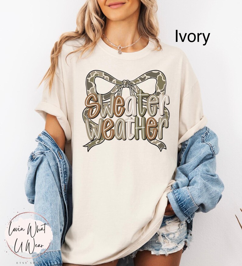 May include: Ivory-colored t-shirt with a bow-shaped graphic in shades of brown and green, featuring the words "Sweater Weather". The shirt is part of a casual wear collection.