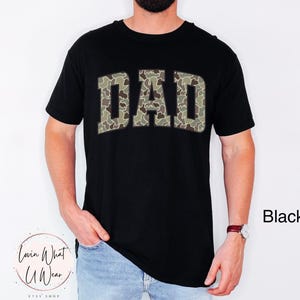 May include: Black crew neck t-shirt with the word "DAD" in a camouflage pattern across the chest. The shirt is worn by a person. The text "Black" is visible in the image.